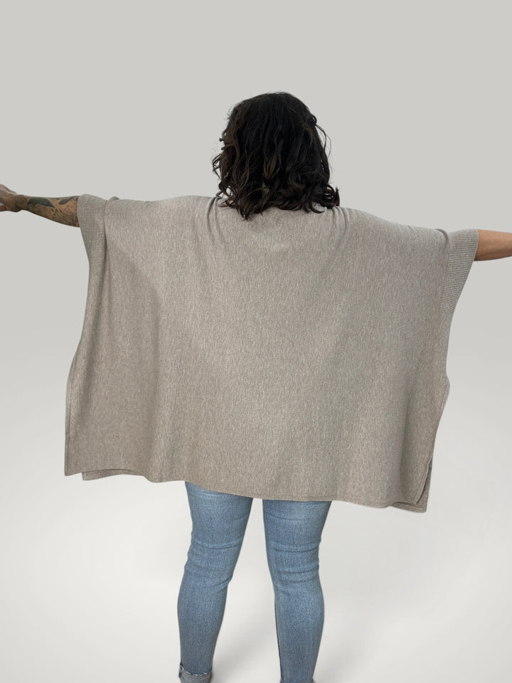 Poncho bege oversized da Isabelle Hannover com gola alta e fendas laterais, visto de costas sobre calças de ganga.