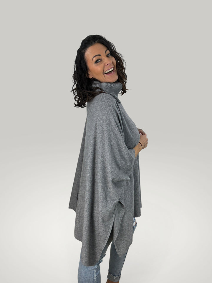 Modelo sorridente veste poncho cinzento Isabelle Hannover com gola alta, corte oversized e mangas curtas em malha