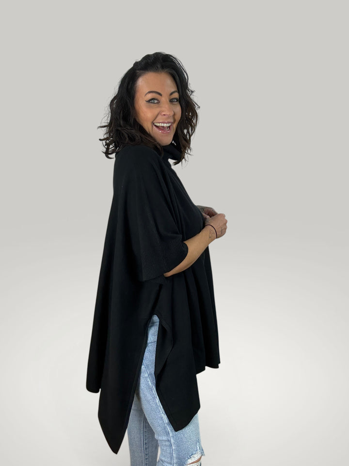 Modelo sorridente veste poncho preto oversized Isabelle Hannover com gola alta e fendas laterais em malha macia